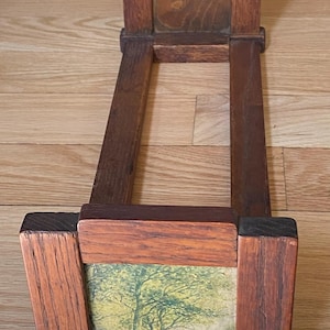 May include: A wooden book rack featuring two upright ends connected by two horizontal rails. The front end displays a rectangular inset panel with a printed landscape illustration of trees in a field, while the back end has a plain wood panel.