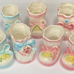 May include: A collection of ceramic baby shoe planters in shades of pink and blue. Each shoe features a decorative bow and floral accents. The planters are designed with a vintage aesthetic.
