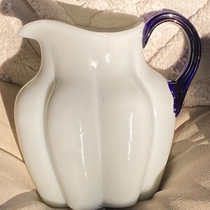 May include: White glass pitcher with a distinctive, ribbed design. The handle is a deep blue color, adding a striking contrast to the white body. The pitcher has a spout for easy pouring and is likely used for serving beverages.