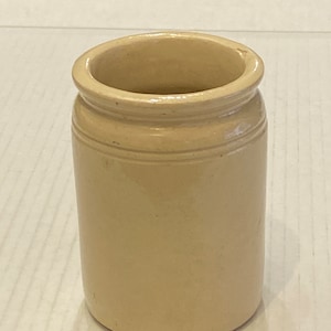 May include: A small, cylindrical, beige stoneware jar with a slightly flared rim. The jar has a smooth, unglazed surface and two horizontal bands near the top. The jar is empty and sits on a white surface.