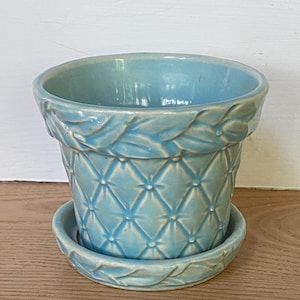 May include: A light blue ceramic plant pot with a matching saucer. The exterior features a raised diamond-shaped quilted pattern with small dots at each intersection and a decorative leaf-like border around the top rim and the saucer edge.