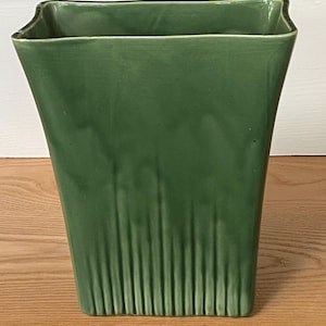 May include: A square-shaped ceramic vase with a glossy forest green glaze. The vessel features a wide, open top that tapers down to a narrower base. The lower portion of the exterior is decorated with vertical, raised ribbing texture.