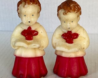 May include: Two vintage candle figurines of children dressed in white robes with red sashes and red skirts, holding songbooks. The candles are made of white wax with painted details.