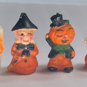 May include: Four Halloween-themed candle figurines. A skull, a witch, a pumpkin, and a ghost. The figurines are made of orange, white, and black wax.