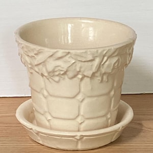 May include: Cream-colored ceramic flower pot with a matching saucer. The pot has a textured, basket-weave design with a decorative leaf pattern around the rim. The saucer is round with a slightly raised edge. Ideal for indoor plants.