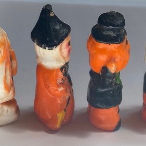 May include: Four Halloween-themed candles, each depicting a different character. The first is a white ghost with orange eyes, the second is an orange witch with a black hat and a white beard, the third is an orange pumpkin with a black hat and a green scarf, and the fourth is a white ghost with a black hat and blue eyes.