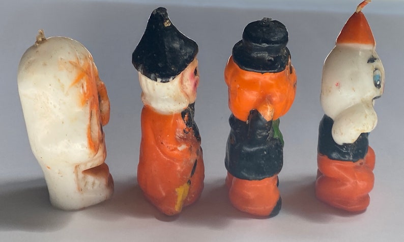 May include: Four Halloween-themed candles, each depicting a different character. The first is a white ghost with orange eyes, the second is an orange witch with a black hat and a white beard, the third is an orange pumpkin with a black hat and a green scarf, and the fourth is a white ghost with a black hat and blue eyes.