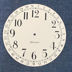 May include: A white clock face with black numerals and a black minute track. The numbers 1 through 12 are displayed, along with the days of the month. The word "Homestead" is printed in cursive.
