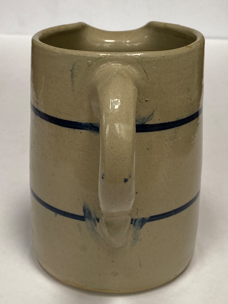 May include: A cream-colored ceramic pitcher with a blue stripe design. The pitcher has a wide mouth and a handle.