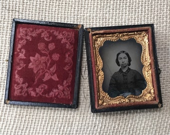Puede incluir: Una fotografía de tintype enmarcada en negro y dorado de una mujer con un vestido oscuro y un cuello blanco. La fotografía está en una caja forrada de terciopelo con un diseño floral.