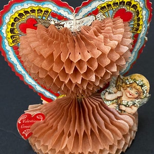 May include: A vintage Valentine's Day card with a honeycomb-shaped paper doll and a cherub holding a bow and arrow. The card features a red heart with the words "To One I Love" and a red heart with the words "Valentine Greetings".