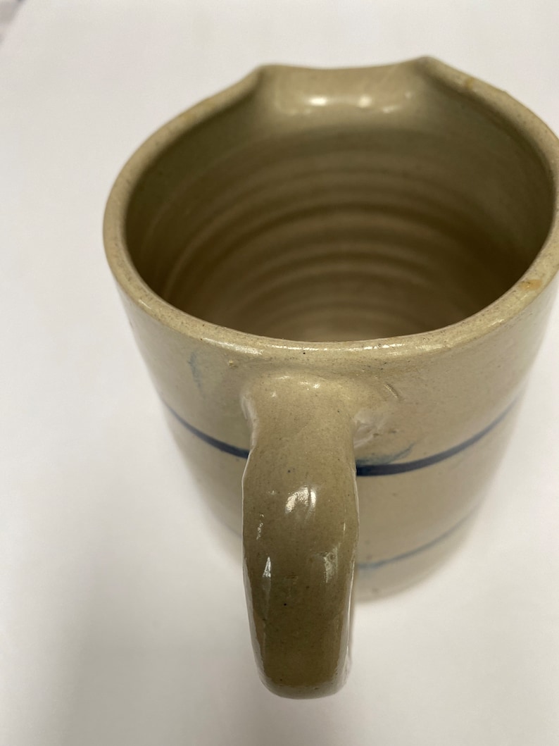 May include: A cream-colored ceramic pitcher with a blue stripe around the middle and a handle. The pitcher has a wide mouth and a rounded bottom.