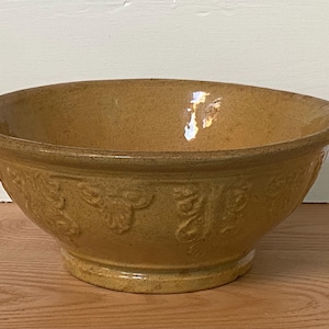 May include: A round, mustard-yellow ceramic mixing bowl with a glossy finish and a wide, flared rim. The exterior surface features a repeating embossed floral or leaf pattern around the middle section, set upon a sturdy, circular base.