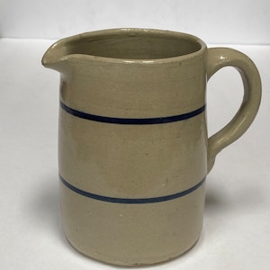 May include: A cream-colored ceramic pitcher with a blue stripe design. The pitcher has a rounded body and a handle.