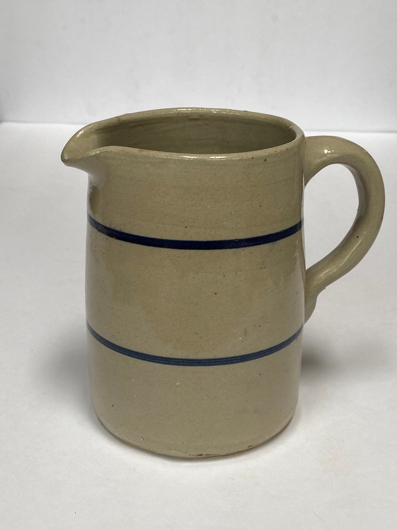 May include: A cream-colored ceramic pitcher with a blue stripe design. The pitcher has a rounded body and a handle.