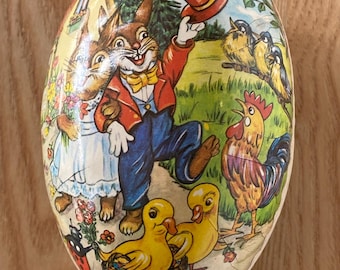 May include: A vintage Easter egg-shaped paper box with a colorful illustration of a bunny wearing a top hat, a rooster, and two ducklings. The bunny is holding a flower bouquet and the ducklings are carrying baskets.