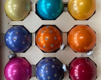 May include: A set of nine colorful Christmas ornaments in a cardboard box. The ornaments are various colors, including gold, teal, orange, blue, and pink. Some have a shiny finish, while others have a speckled design. Each ornament has a silver metal cap.