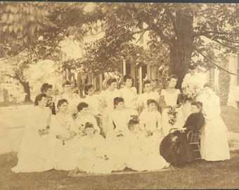 May include: Sepia-toned photograph of a group of women in white dresses posing outdoors. The women are arranged on a lawn in front of a large tree and a house. The image captures a moment in time, likely a formal gathering.