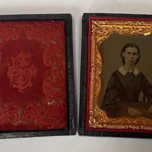 May include: An antique portrait in a black case. The portrait features a woman in a dark dress with a white collar, set within a gold-colored ornate frame. The case is open, revealing a red velvet interior with an embossed design.