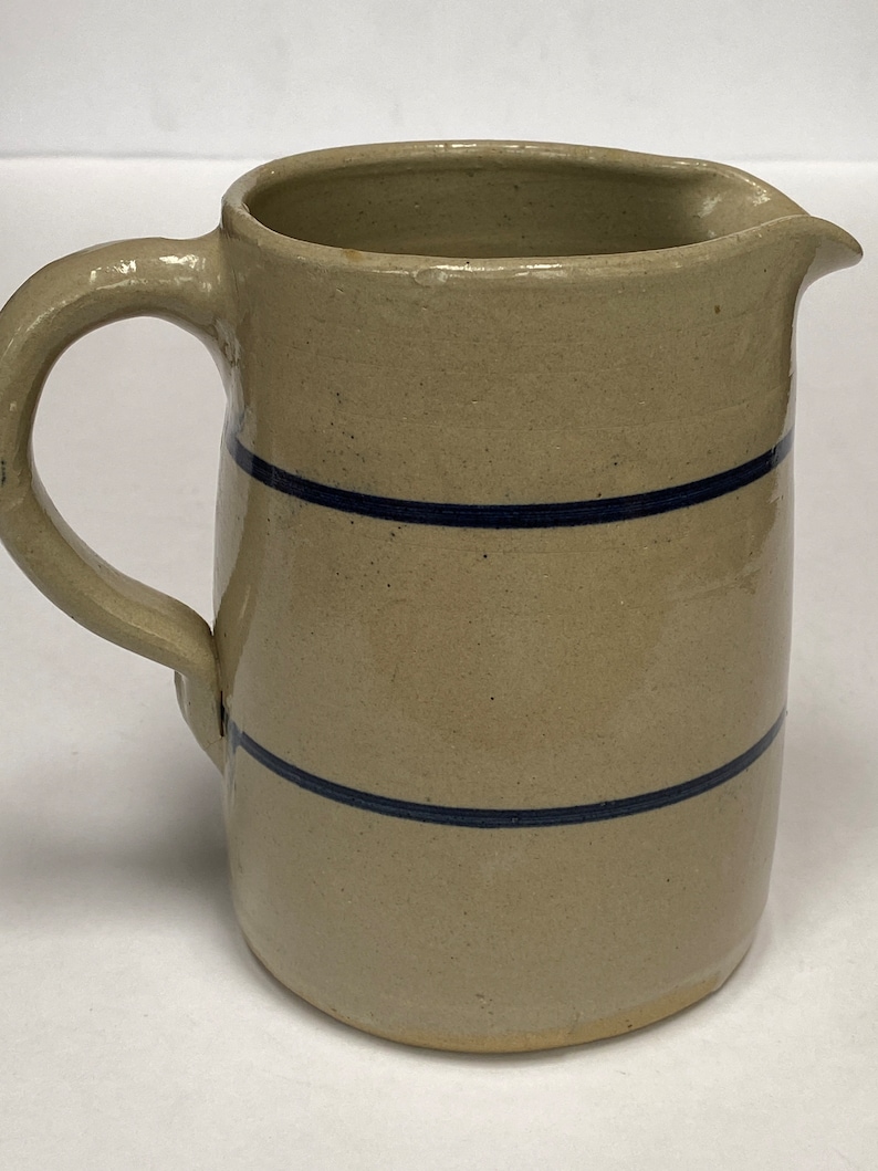 May include: A beige stoneware pitcher with two blue bands around the body. The pitcher has a rounded body and a handle.