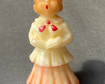 May include: A vintage candle shaped like a person singing, with a white dress and a red bow. The candle has a light brown complexion and a red mouth.