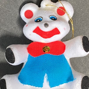 May include: White plush bear ornament with red ears, a red bow tie, and a blue vest. The bear has black eyes and a red mouth.