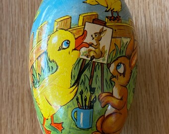 May include: A vintage Easter egg-shaped cardboard container with a colorful illustration of a duck, a rabbit, and a chick. The duck is holding a paintbrush and pointing to a painting of a rabbit on an easel. The rabbit is sitting on the ground with a paintbrush in its paw. The chick is perched on a fence. The container has a blue, green, and yellow color scheme.