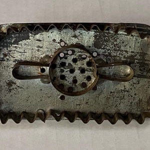 May include: A vintage, rectangular metal cookie cutter with a decorative, scalloped edge. The center features a circular design with small holes. The metal has a worn, aged appearance, suggesting it is an antique kitchen tool.