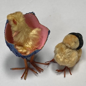 May include: Two vintage, miniature, plush chicks. One chick is inside a blue and pink eggshell with orange legs. The other chick is yellow with a black hat and orange legs. These are likely Easter decorations.