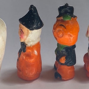 May include: Four Halloween-themed candle figurines. The figurines are made of wax and are painted in orange, black, and white. The figurines depict a witch, a pumpkin, a ghost, and a skeleton.