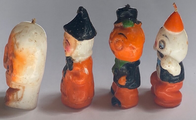 May include: Four Halloween-themed candle figurines. The figurines are made of wax and are painted in orange, black, and white. The figurines depict a witch, a pumpkin, a ghost, and a skeleton.