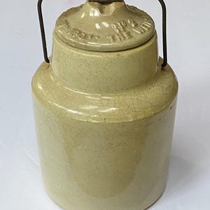 May include: A vintage, light-yellow stoneware crock with a lid and a metal handle. The lid is embossed with text, and the crock has a cylindrical shape. The overall design is simple and rustic, suggesting a piece of antique kitchenware.