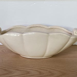 May include: Cream-colored, scalloped-edge ceramic bowl with two scrolled handles. The bowl has a wide, shallow design and sits on a wooden surface. The bowl's shape resembles a seashell.