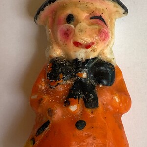 May include: A vintage Halloween candle shaped like a witch. The witch is wearing a black hat and an orange dress with black polka dots. The candle has a whimsical expression with one eye closed and a slight smile.