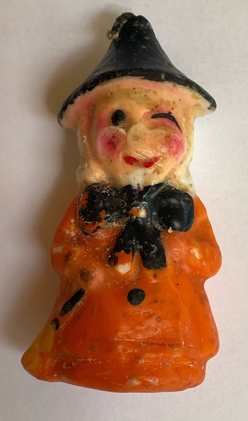 May include: A vintage Halloween candle shaped like a witch. The witch is wearing a black hat and an orange dress with black polka dots. The candle has a whimsical expression with one eye closed and a slight smile.