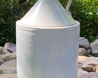May include: A white ceramic crock with a rounded body and a handle. The crock is sitting on a pile of rocks.