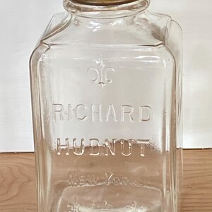 May include: A clear glass jar with a gold metal lid. The jar is labeled "Richard Hudnut" and has a decorative design on the front.