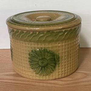 May include: A cylindrical ceramic canister with a lid, featuring a tan waffle-grid texture and a raised green daisy motif on the side. The rim is decorated with a green wavy pattern, and the lid has a matching green border and a central knob.
