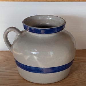 May include: A white ceramic pitcher with a blue rim and two blue bands around the body. The pitcher has a handle and is a traditional style.