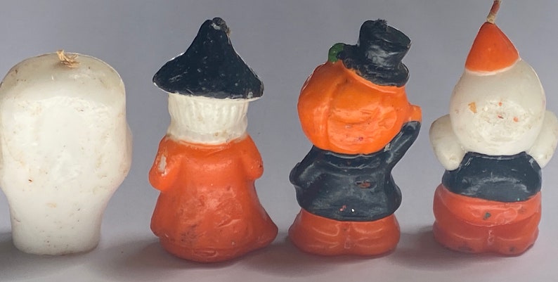 May include: Four Halloween-themed candle figurines. A white ghost, a witch with a black hat and orange dress, a pumpkin with a black hat and orange face, and a white ghost with a red hat and orange trousers.