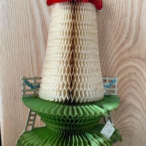 May include: A vintage paper craft in the shape of a mushroom with a red top, white stem, and green base. The craft is made of honeycomb paper and has a delicate, intricate design. The base of the mushroom has a small fence and a ladder.