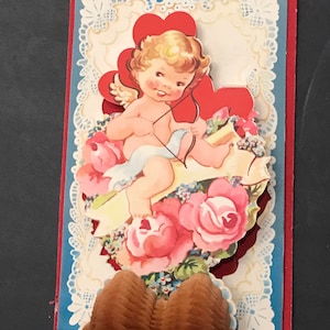 May include: A vintage Valentine's Day card with a pop-up design. A cherub with wings and a bow and arrow sits on a cloud with pink roses. The card has a blue and white lace border.