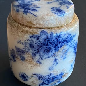 May include: A white ceramic jar with a lid, featuring blue floral patterns. The jar has a cylindrical shape with a gold-colored band around the top and bottom. The lid also has a gold band and blue floral designs.
