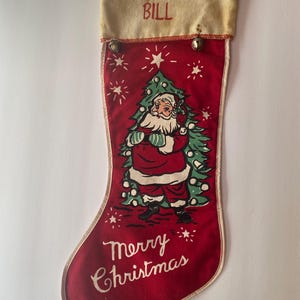 May include: A red Christmas stocking featuring a graphic of Santa Claus standing in front of a green pine tree. The white cuff is embroidered with the name BILL in red thread. The text Merry Christmas is printed in white at the bottom.