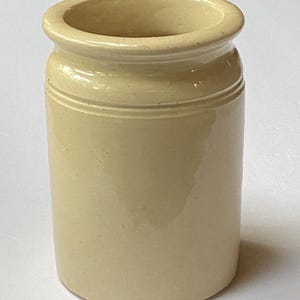 May include: A small, cylindrical, stoneware jar with a light beige glaze. The jar has a rounded rim and two horizontal bands near the top. The overall shape is simple and classic, suitable for various uses.