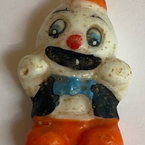 May include: A vintage clown candle with a white face, blue eyes, a red nose, and a black and blue bow tie. The clown is wearing an orange hat and orange trousers.