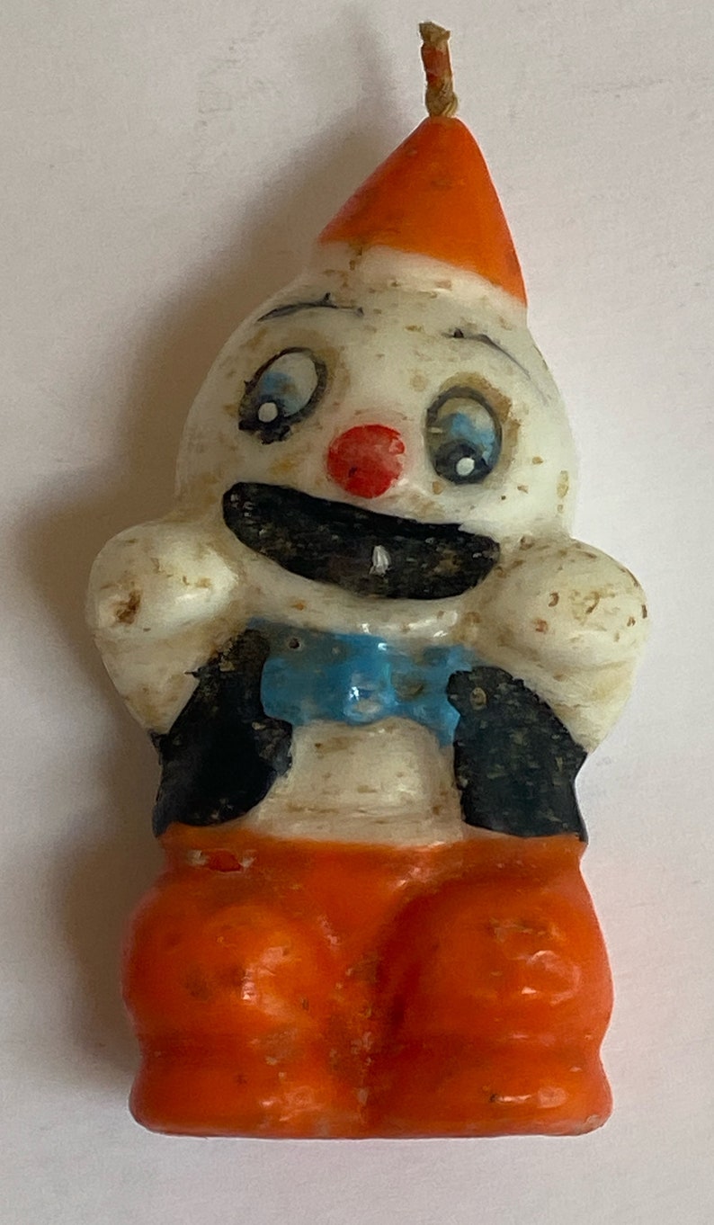 May include: A vintage clown candle with a white face, blue eyes, a red nose, and a black and blue bow tie. The clown is wearing an orange hat and orange trousers.