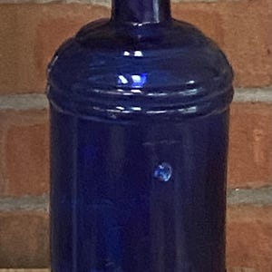 May include: A cobalt blue glass bottle with a cylindrical body and a narrow neck. The bottle has a rounded shoulder and base, with horizontal ridges. The text "S. STAFFORD" and "MADE IN USA" is embossed on the bottle.