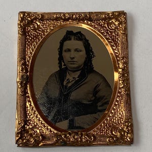 May include: An antique portrait of a woman in a gold-colored ornate frame. The oval portrait is sepia-toned, showing a woman with dark hair and a dark jacket. The frame has a detailed, embossed design.