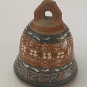 May include: A ceramic bell with a brown, gray, and white design. The bell has a loop at the top and is decorated with spiral and floral patterns. The base of the bell has a geometric design.